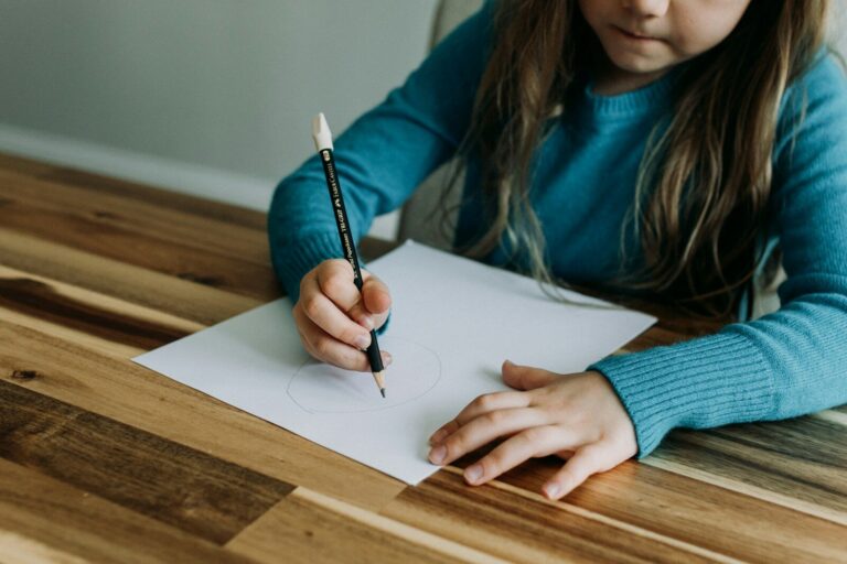 Home 12 Young child holding a pencil and writing on paper at a table, showing pencil grip and fine motor skills
