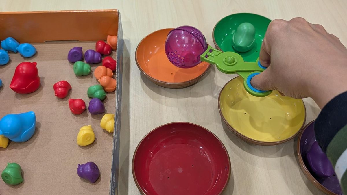 Hand using a squeeze scoop to sort coloured toy birds into matching bowls, a fine motor play activity