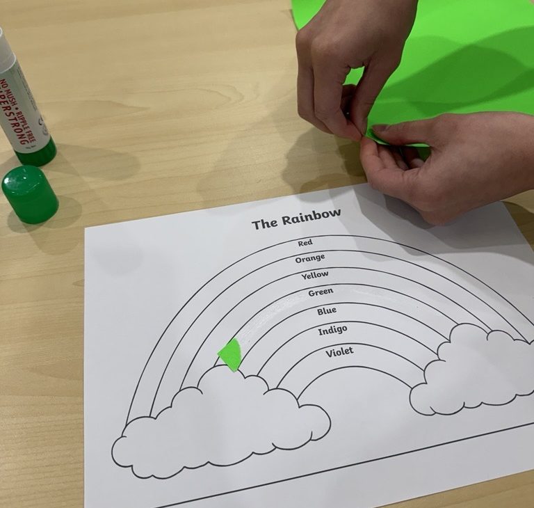 Torn green paper being glued onto the green band of a rainbow worksheet next to a glue stick