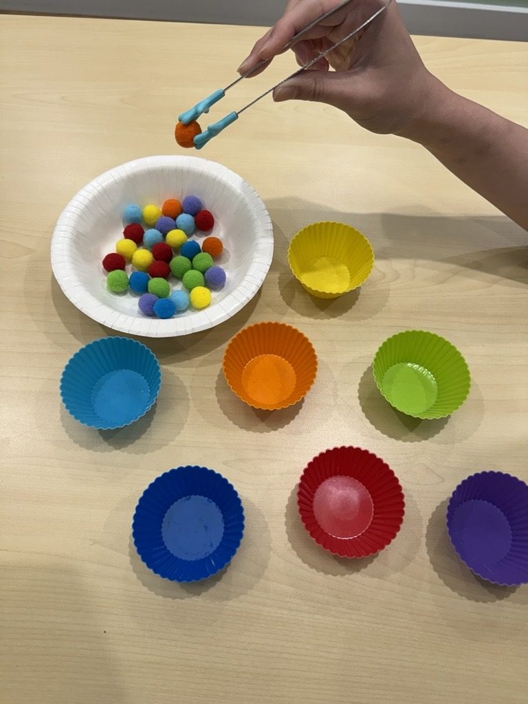 Lifting an orange pom-pom with tongs using a pinch grasp, ready to drop it into the orange cup