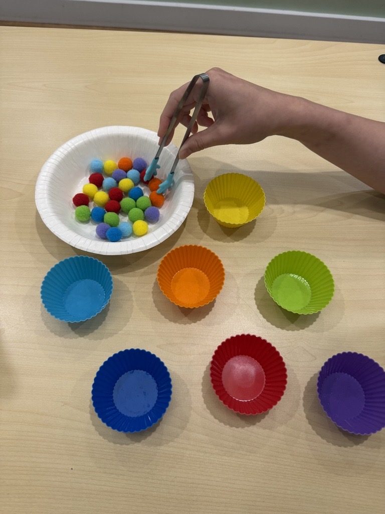 Tongs picking up pom-poms from a bowl to sort into rainbow-coloured silicone cups